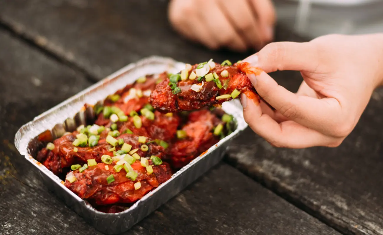 Hand picking up a spicy chicken wing garnished with chopped green onions from a metal tray on a wooden table
