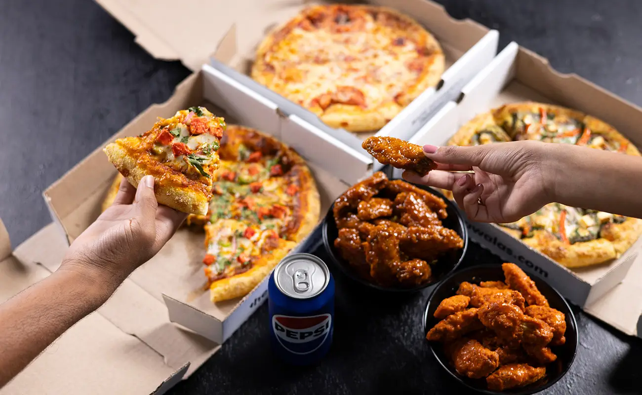 Two open pizza boxes with veggie and cheese pizzas, bowls of saucy chicken wings, a can of Pepsi, and hands holding a pizza slice and a wing on a dark surface