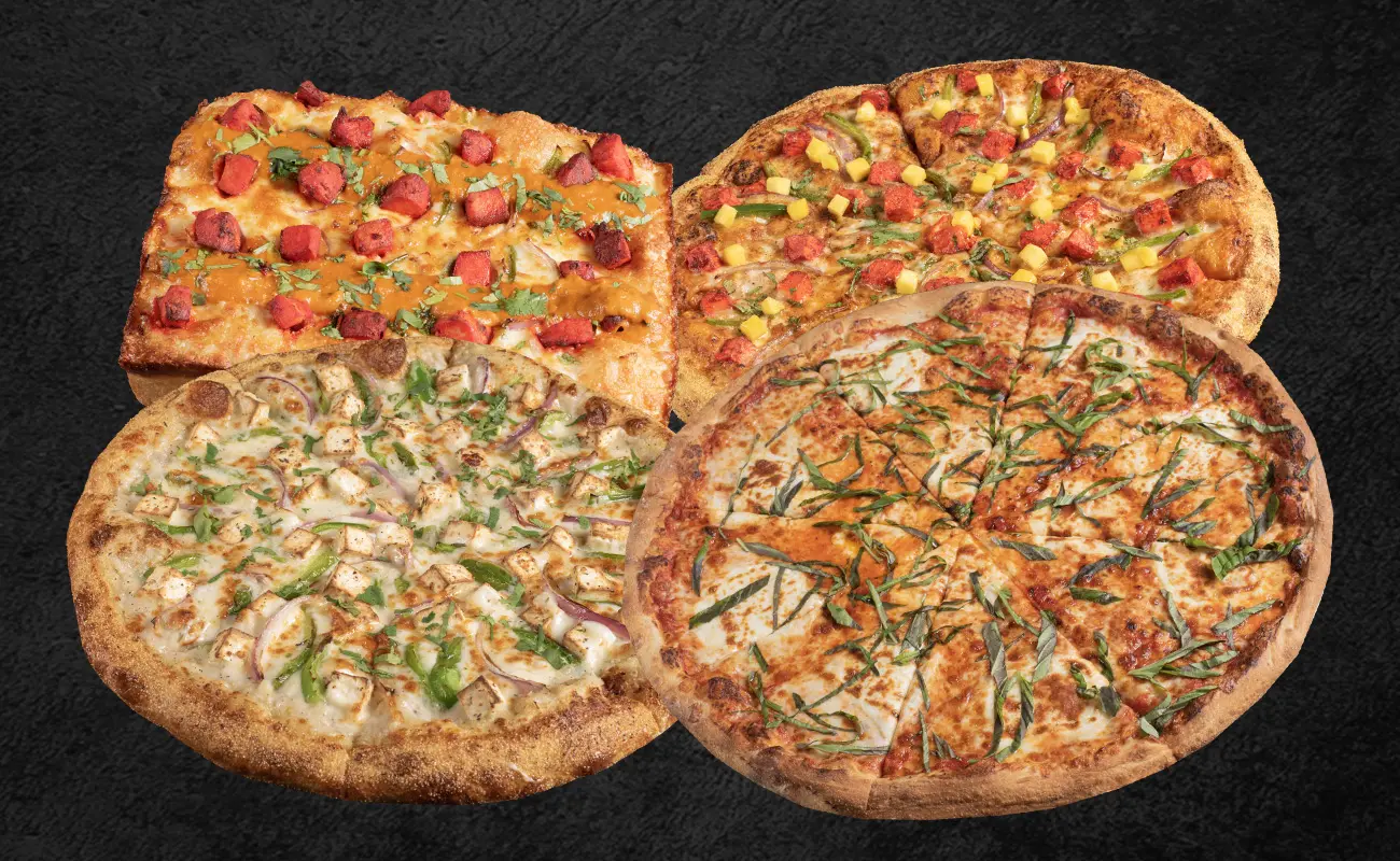 An assortment of four gourmet pizzas displayed on a dark textured background. The pizzas feature a variety of toppings including paneer cubes, chopped cilantro, sliced onions, bell peppers, cheese, tandoori-style toppings, and fresh basil. One of the pizzas is rectangular, while the others are round.