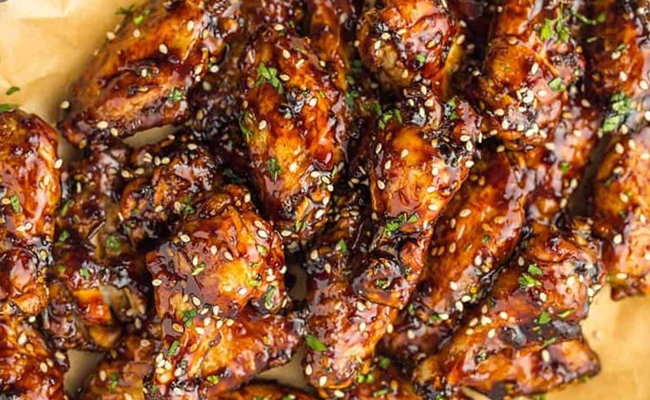 This image is a close-up of dark, richly glazed chicken wings generously covered in sesame seeds and chopped parsley.